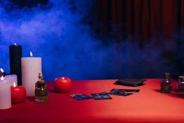 KYIV, UKRAINE - JUNE 29, 2022: burning candles and tarot cards on red table near blue smoke on black background