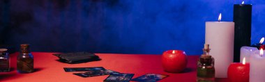 KYIV, UKRAINE - JUNE 29, 2022: essential oils and tarot cards near burning candles on red table near blue smoke on dark background, banner