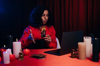 KYIV, UKRAINE - JUNE 29, 2022: brunette oracle holding tarot card during online esoteric session on laptop on black background with red drape
