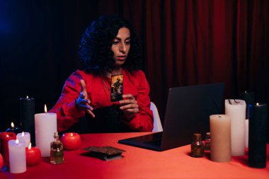KYIV, UKRAINE - JUNE 29, 2022: brunette oracle holding tarot card during online esoteric session on laptop on black background with red drape clipart
