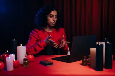 brunette future teller pointing at laptop during online spiritual session on dark background
