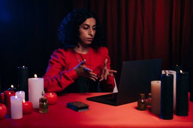 brunette future teller pointing at laptop during online spiritual session on dark background clipart