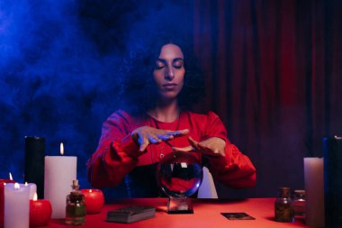soothsayer with closed eyes near crystal ball and burning candles on dark background with blue smoke