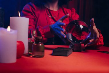 cropped view of fortune teller near crystal ball and burning candles on dark background