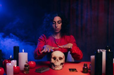 brunette oracle during magic session near skull and burning candles on dark background with blue smoke