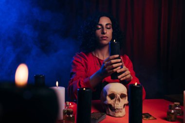 oracle with closed eyes holding burning candle near skull and blue smoke on dark background