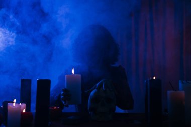 medium holding candle and touching skull during spiritual session in darkness with blue smoke
