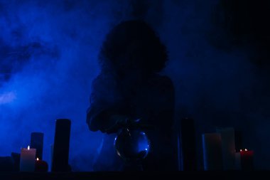 fortune teller holding hand above crystal ball near burning candles in darkness with blue smoke