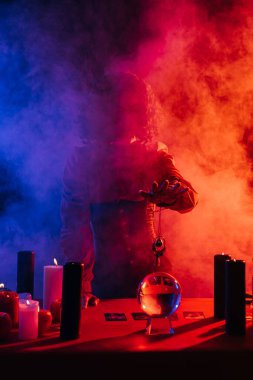 fortune teller holding amulet near crystal ball and candles near colorful smoke on foreground
