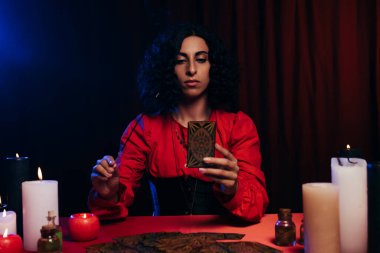 KYIV, UKRAINE - JUNE 29, 2022: brunette medium with palo santo stick and tarot card near burning candles on dark background