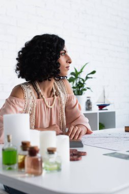 young astrologist looking away near candles and bottles with essential oils 