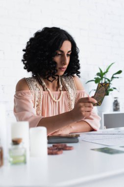 KYIV, UKRAINE - JUNE 29, 2022: young oracle looking at tarot card near blurred candles