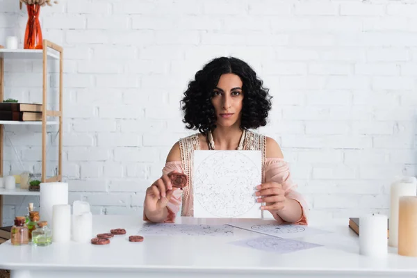 brunette astrologist showing constellations map and clay rune while looking at camera