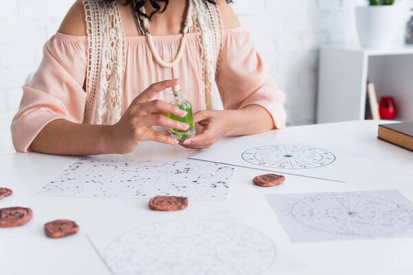 partial view of fortune teller holding bottle of essential oil near blurred clay runes and natal charts 