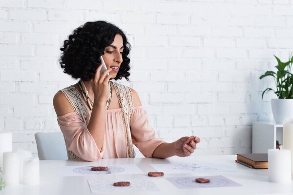 smiling astrologer holding clay rune while talking on smartphone at home