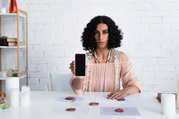 brunette fortune teller looking at camera while holding smartphone with blank screen near clay runes and star maps