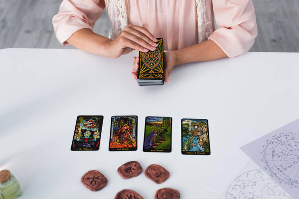 KYIV, UKRAINE - JUNE 29, 2022: partial view of predictor holding tarot cards near clay runes during spiritual session