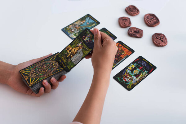 KYIV, UKRAINE - JUNE 29, 2022: high angle view of cropped soothsayer holding tarot cards near blurred clay runes