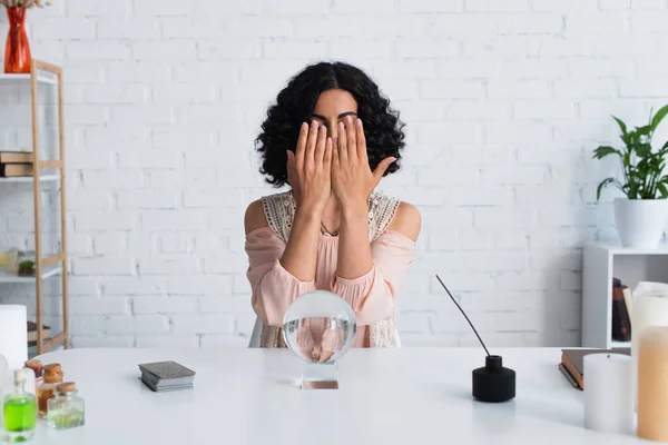 brunette fortune teller obscuring face with hands near magic orb and diffuser with aroma stick