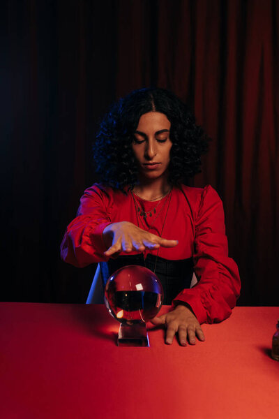 brunette fortune teller with closed eyes holding hand over crystal ball on dark background with red drape