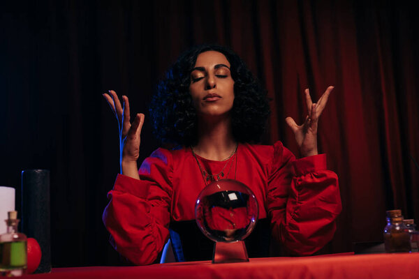 medium with closed eyes sitting near crystal ball on dark background