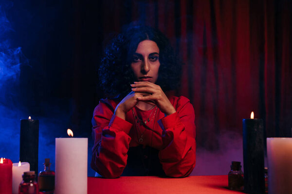 young medium sitting near burning candles on dark background with blue smoke and red drape
