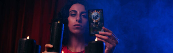KYIV, UKRAINE - JUNE 29, 2022: young oracle with tarot card and burning candle near blue smoke on dark background, banner