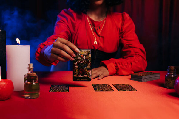 KYIV, UKRAINE - JUNE 29, 2022: cropped view of medium with tarot card near essential oils and burning candles on dark background