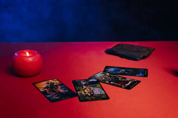 KYIV, UKRAINE - JUNE 29, 2022: tarot cards and burning candle on red table and black background with blue smoke