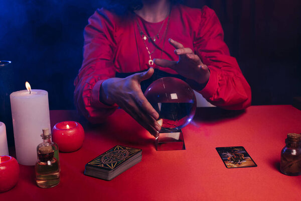 KYIV, UKRAINE - JUNE 29, 2022: partial view of oracle with crystal ball near tarot cards and burning candles on dark background