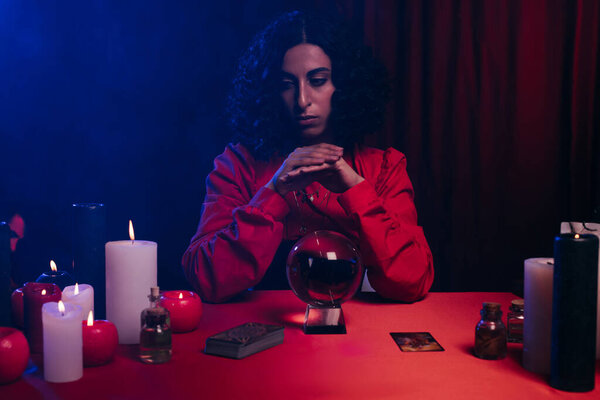 KYIV, UKRAINE - JUNE 29, 2022: brunette medium near crystal ball and burning candles on dark background