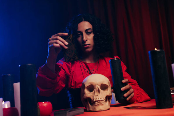 young medium sitting near skull and burning candles on dark background with red drapery