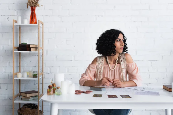 brunette fortune teller looking away while sitting near tarot cards and cosmic charts