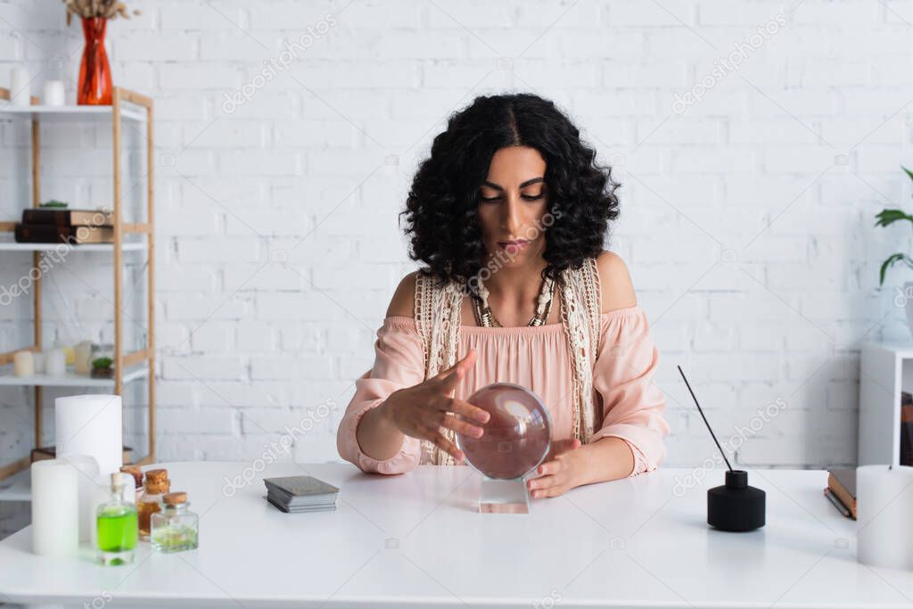 brunette astrologist touching magic sphere near tarot cards and diffuser with aroma stick