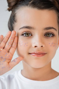 portrait of adorable girl with lip gloss and sparkling glitter stars on face looking at camera isolated on grey clipart