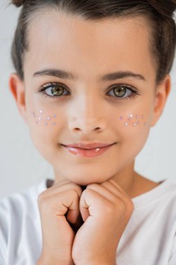 portrait of happy girl with lip gloss and sparkling glitter stars on face looking at camera isolated on grey