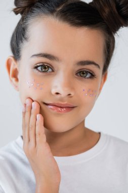 portrait of smiling girl with lip gloss and sparkling glitter stars on face touching cheek isolated on grey