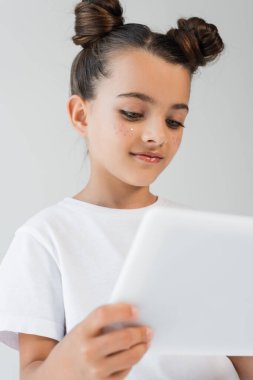 cheerful girl with glitter stars on cheeks smiling while using digital tablet isolated on grey