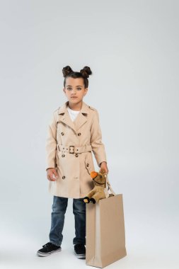 full length of stylish child in trench coat and jeans holding shopping bag with toy horse on grey