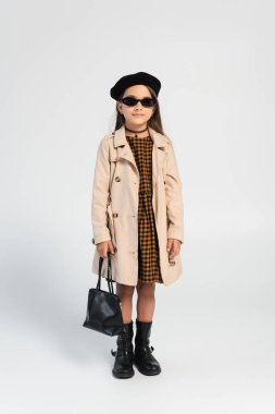 full length of stylish girl in trench coat and beret holding handbag on grey