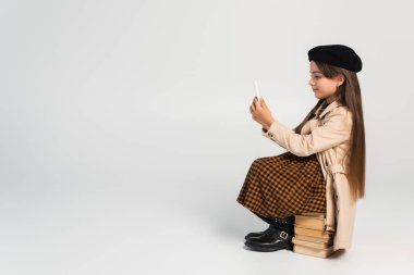 full length of cute kid in stylish autumnal outfit and beret sitting on stacked books and using smartphone on grey
