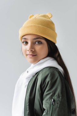 portrait of adorable girl in yellow beanie hat and bomber jacket looking at camera isolated on grey clipart