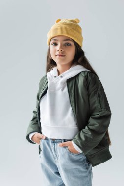 portrait of adorable girl in yellow beanie hat and stylish autumnal outfit posing with hands in pockets isolated on grey  clipart