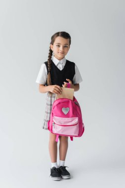 full length of cute schoolgirl in uniform holding pink backpack and book on grey 