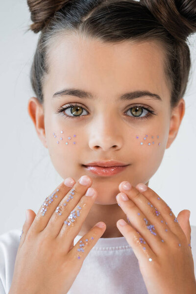 portrait of adorable girl with sparkling glitter stars on face and hands looking at camera isolated on grey