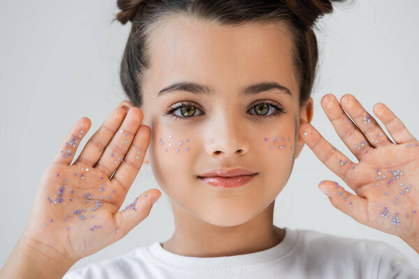 portrait of adorable girl with sparkling glitter stars on face and palm hands looking at camera isolated on grey