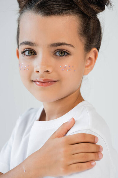 portrait of cheerful girl with lip gloss and sparkling glitter stars on face looking at camera isolated on grey