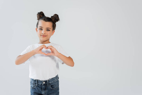 cheerful girl with sparkling glitter stars on cheeks showing heart with hands isolated on grey