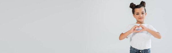 cheerful girl with  glitter stars on cheeks showing heart with hands isolated on grey, banner