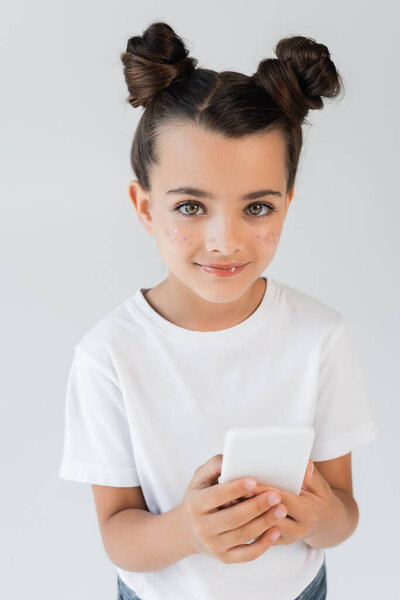 adorable girl with glitter stars on cheeks smiling while using smartphone isolated on grey